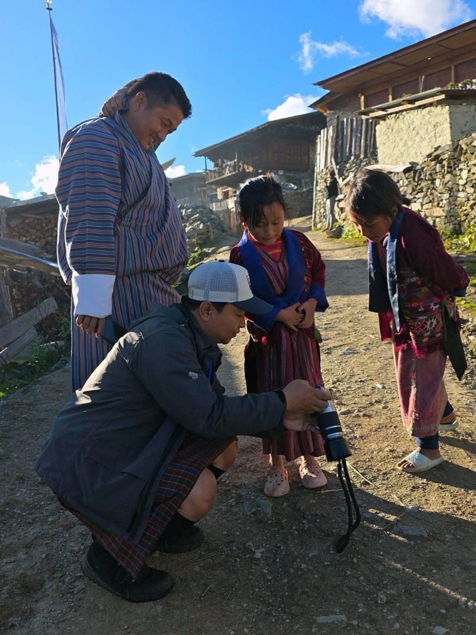 A guide in a cap crouches on a village path to show two children a photo on a camera, while another guide in a gho looks on