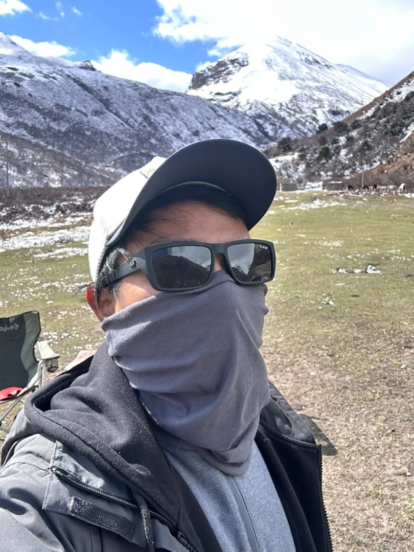 Tashi in a cap, sunglasses and a grey neck gaiter, with snow-dusted mountains and a grassy valley behind